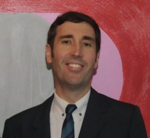 Colored photograph of head and shoulders of smiling man with dark hair, wearing dark suit, white shirt and dark tie. Background painted in 3 colors grey , pale pink and dark pink.