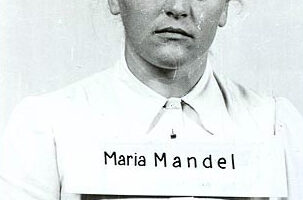 Black and White photograph of female prisoner in white blouse with name banner attached and the words "Maria Mandel" printed on it. Female Perpetrators of the Holocaust.